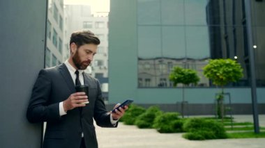 Molada resmi takım elbiseli sakallı iş adamı cep telefonu kullanıyor. Şehir merkezindeki modern şehir sokaklarının arka planında elinde kahve fincanıyla duran bir iş adamı. Boşluğu kopyala