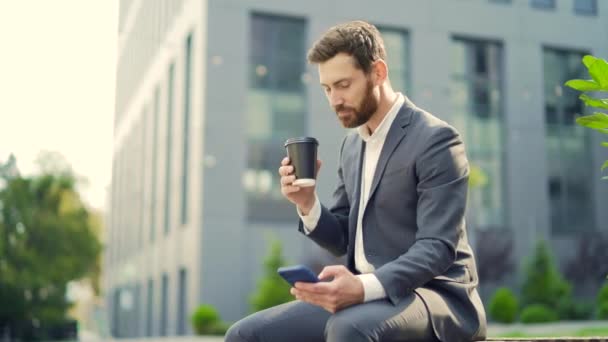 カップコーヒーとベンチに座ってハンサムな幸せなひげを生やしたビジネスマンは 携帯電話を使用してスマートフォンを手で閲覧良いニュースと喜びの笑顔を読み取ります 近代的な都市の通りの背景の外 シティパーク — ストック動画