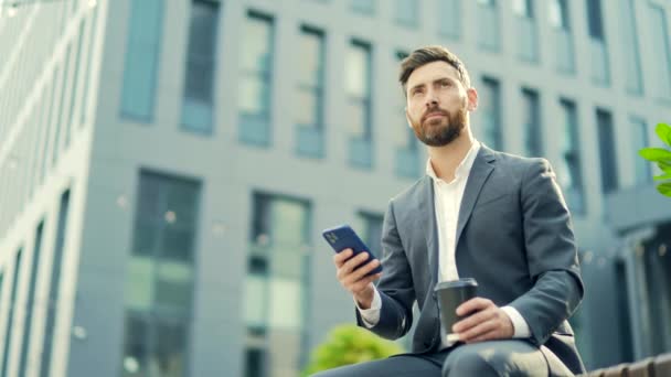 コーヒーを飲みながらベンチに座っているハンサムな髭のビジネスマンは 携帯電話を使用してスマートフォンを手でブラウズし スマートを使用するというニュースを読み取ります ダウンタウンだ 近代的な都市の通りの背景の外 シティパーク — ストック動画