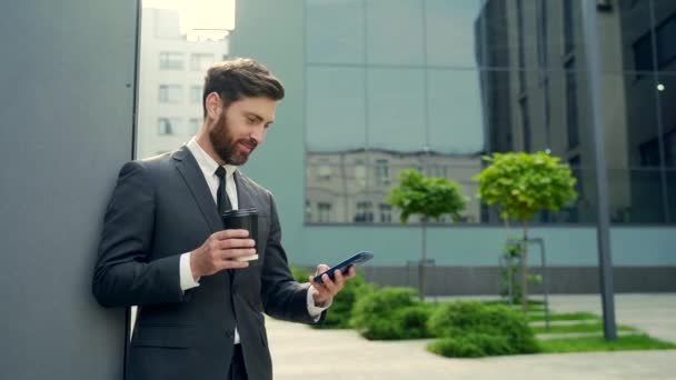 戴胡子的商人在休息时用智能手机穿正装 商人站在现代城市街道的背景上 在市中心的户外端着咖啡杯 复制空间 — 图库视频影像