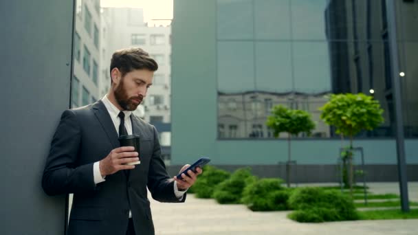 携帯電話の使用のスマートフォンを使用して休憩中に正式なスーツで髭ビジネスマン 郊外のダウンタウンにコーヒーカップを持つ現代の都市の通りの背景に立っているビジネスマン コピースペース — ストック動画
