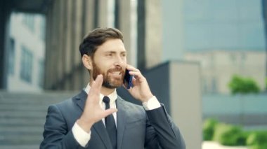 Başarılı genç iş adamı arka planda şehir merkezindeki modern ofis binasından bahsediyor. Sakallı iş adamı resmi takım elbise giymiş bir cep telefonu açık şehir caddesinde duruyor.