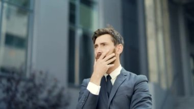 Üzücü bir iş adamı sakallı, beyaz bir adam kafasını tutuyor. İş adamı, girişimci, işçi ya da takım elbiseli bir işçi modern ofis merkezinin arka planında. Dışarı çıkın. Açık havada.