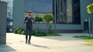 Kendine güvenen başarılı genç işadamı cep telefonuyla şehir merkezindeki modern ofis binasına karşı hareket ediyor. İş adamı resmi bir takım elbise içinde kahve fincanı kullanıyor ve yürüyor.