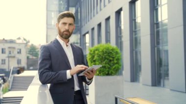 Resmi takım elbiseli şık sakallı iş adamı dışarıda arka plandaki modern ofis binasında elinde tabletle çalışıyor. Adam akıllı telefon kullanıyor ya da şehir dışında cep telefonu kullanıyor.