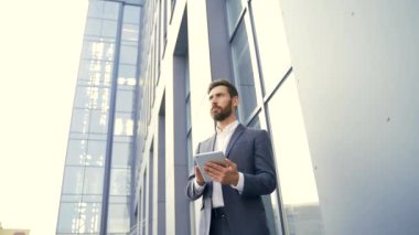 Resmi takım elbiseli şık sakallı iş adamı dışarıda arka plandaki modern ofis binasında elinde tabletle çalışıyor. Adam akıllı telefon kullanıyor ya da şehir dışında cep telefonu kullanıyor.