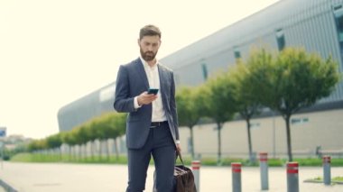Seyahat çantası ve cep telefonu taşıyan şık sakallı bir iş adamı şehir sokaklarında modern şehir geçmişinde yürüyor. Takım elbiseli bir iş adamı akıllı telefon kullanır. Turist gezisi. Kafkas Yöneticisi