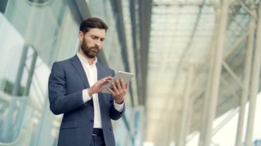Resmi takım elbiseli şık sakallı iş adamı dışarıda arka plandaki modern ofis binasında elinde tabletle çalışıyor. Adam akıllı telefon kullanıyor ya da şehir dışında cep telefonu kullanıyor.