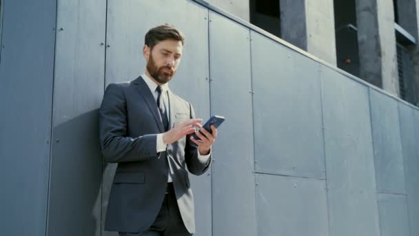 都市部のバックグラウンドオフィスのテキストメッセージを構築する白人ひげを生やした従業員 良いニュース携帯電話を読む ビジネスマンは屋外でスマートフォンの携帯電話を使用して正式なスーツメッセージを立って — ストック動画