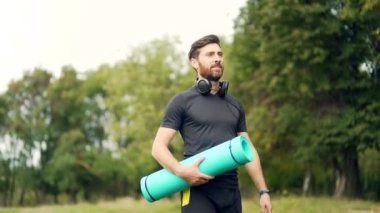 Aktif yakışıklı erkek yogi antrenmandan önce ya da sonra parkta dolaşır. Genç sakallı atlet, elinde spor minderiyle yoga sınıfına gidiyor. Açık hava Sağlıklı yaşam tarzı