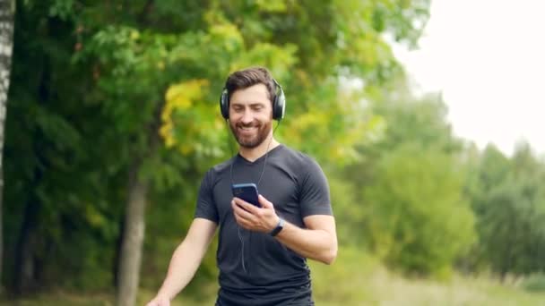 年轻活泼活泼的小伙子 带着耳机散步 喜欢听音乐 在城市公园或森林背景下慢跑 男人做健身锻炼耳机 — 图库视频影像