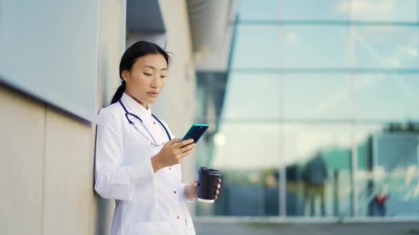 在医院门诊部的墙上 一位年轻的亚洲女医生或助理医生在休息时精疲力尽地站着 手里拿着一杯咖啡和一部电话 实习生在外面用智能手机 — 图库视频影像