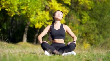 Genç, güzel spor kızı Asyalı kadın parkta meditasyon yapıyor, lotus pozu veriyor yoga minderi yapıyor, zen. Sabahları doğada rahatlar. Sağlıklı yaşam tarzını kavrayın, rahatlayın, sakinleşin, meditasyon yapın.