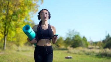 Aktif güzel Asyalı dişi yogi antrenmandan önce ya da sonra parkta dolaşır. Genç atletik formda bir kadın yoga kursuna spor minderiyle gider, kulaklıkla müzik dinler. Açık havada.