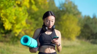 Aktif güzel Asyalı dişi yogi antrenmandan önce ya da sonra parkta dolaşır. Genç atletik formda bir kadın yoga kursuna spor minderiyle gider, kulaklıkla müzik dinler. Açık havada.