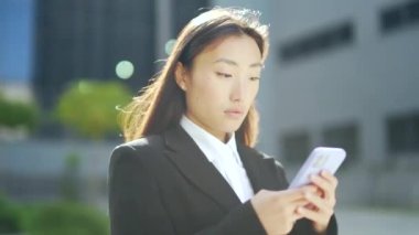 Asyalı bir çalışan şehir merkezindeki modern ofis binasının arka planında cep telefonuna mesaj atıyor. İş kadını resmi bir takım elbiseyle şehrin dışındaki akıllı telefondan mesaj atıyor.