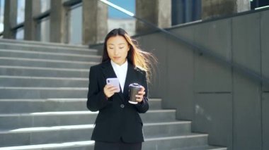 Kendine güvenen bir yönetici olan iş kadını, büyük bir modern iş bölgesinin yakınında kentsel bir caddede duruyor. Genç Asyalı kadın Smartphone 'da mesajlar yolluyor ve elinde bir kahve fincanı tutuyor. Başarı 