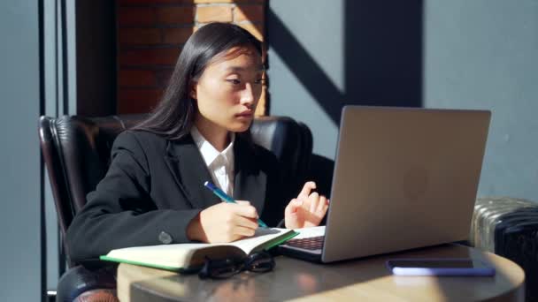 年轻的女自由职业者或在网上工作或在咖啡馆远程学习的学生 身穿西服的亚洲女商人在工作场所的办公桌旁使用笔记本电脑打字 写在笔记本上 — 图库视频影像