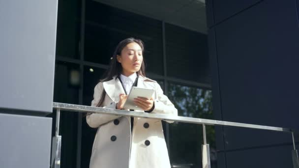 テラスの手すり近くに立つ若いアジア系女性のビジネスマンデジタルタブレットを使ってモダンなオフィスビル 街の通りに都市部の背景に成功したビジネス女性 — ストック動画
