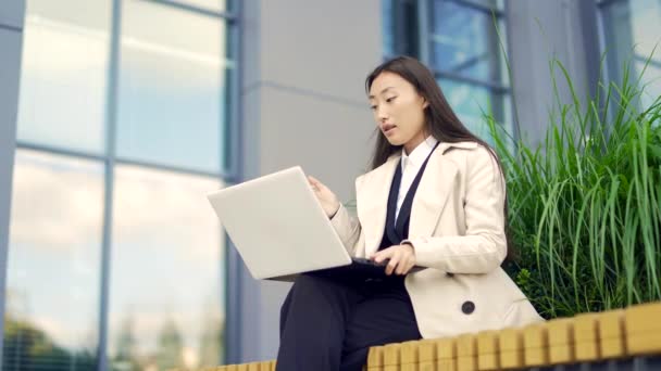 現代のオフィスセンターの都市を背景にベンチに座ってリモートで働く若いアジアのビジネス女性 ビジネスマンは ラップトップ会議を使用してオンラインでビデオ通話を話しています 外部との通信 — ストック動画