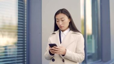 Asyalı bir çalışan şehir merkezindeki modern ofis binasının arka planında cep telefonuna mesaj atıyor. İş kadını resmi bir takım elbiseyle şehrin dışındaki akıllı telefondan mesaj atıyor.