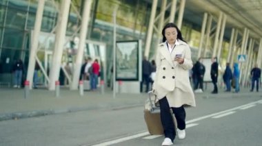 Asyalı kadın turist iş kadını terminal havaalanından bavuluyla yürüyor. Şehir merkezindeki modern bir tren istasyonu. Elinde cep telefonu tutarak. Kadın iş gezisi