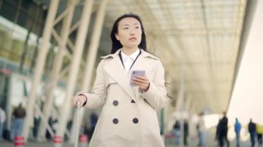Arka planda duran Asyalı genç iş kadını portresi. Paltolu modern tren istasyonu havaalanı ve uygulama cep telefonu kullanıyor. Seyahat eden iş kadını turist ellerinde akıllı telefon kullanıyor.