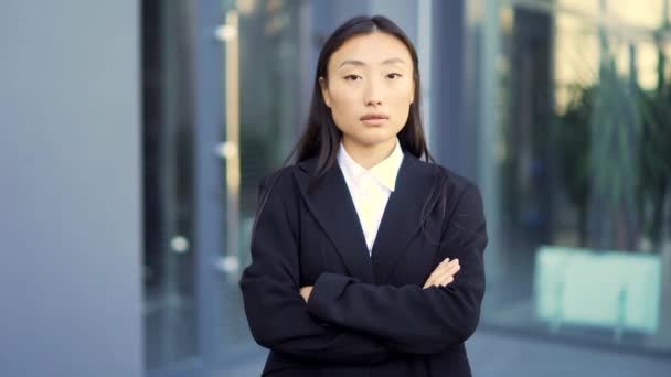 若い自信に満ちたアジアの実業家がカメラを見ています 外の近代的なオフィスビルの暗い都市部の背景に立って深刻なビジネス女性 女性の正式なスーツの腕が交差 — ストック動画