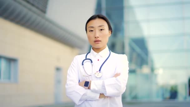 近照亚洲女医生的肖像 在外面的现代医院的背景下 手挽手看着相机 自信的医生在室外 女科学家治疗师在诊所附近穿着洗涤剂 — 图库视频影像