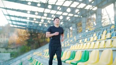 Genç Asyalı koşucu modern şehir stadyumunun arka planında koşuyor. Siyah spor kıyafetleri içinde sabah koşusu sporu yapan bir adam. Şehir caddesinde koşuyor. Açık hava köprüsü. Ağır çekim. Sağlıklı Yaşam Tarzı