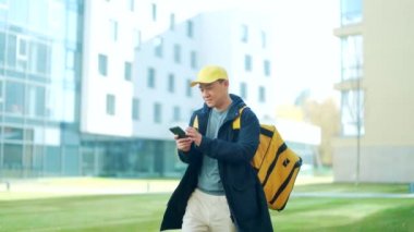 Sarı üniformalı Asyalı erkek yemek kuryesi akıllı telefondan cep telefonu uygulamasını kullanarak şehir caddesinde izole bir şekilde yürürken termal sırt çantasıyla yiyecek dağıtıyordu. Müşteri adresine bakıyorum. çevrimiçi satın al