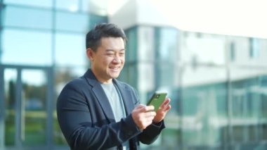 Neşeli Asyalı adam modern şehir binasının önünde akıllı telefon kullanmayı seviyor. Komik haberler okuyor. Erkek gülümsemesi kayıyor, daktilo ediyor, cep telefonu kullanıyor. İnternetten sipariş ya da alım yapar