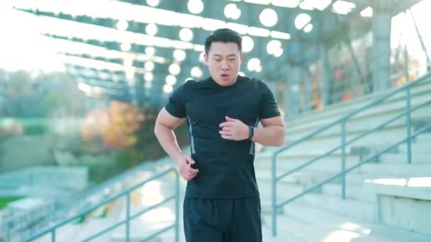 体育亚洲人在现代田径上看起来很聪明 男人跑步用聪明的手镯 人物形象健康检查结果 男子穿着运动服在城市街道的后台体育场慢跑 — 图库视频影像