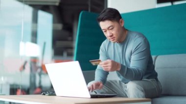 Mutlu Asyalı adam internetten alışveriş yapıyor. Kredi kartıyla ödeme yapan müşteri. Modern bir ofis yerinde oturuyor. Seyahat, servis, rezervasyon. Serbest çalışan, serbest çalışan bir erkek. Bir dizüstü bilgisayar kullanarak