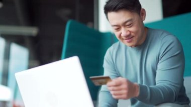 Mutlu Asyalı adam internetten alışveriş yapıyor. Kredi kartıyla ödeme yapan müşteri. Modern bir ofis yerinde oturuyor. Seyahat, servis, rezervasyon. Serbest çalışan, serbest çalışan bir erkek. Bir dizüstü bilgisayar kullanarak