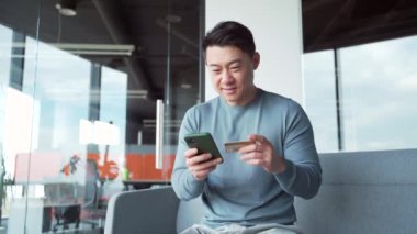 Mutlu Asyalı adam internetten alışveriş yapıyor. Modern bir ofiste banka kartıyla ödeme yapan bir müşteri. Seyahat, servis, rezervasyon. Serbest çalışan bir erkek, cep telefonu akıllı telefonu kullanıyor.