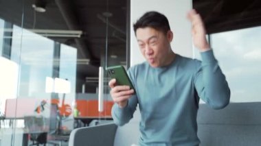 Mutlu Asyalı serbest çalışan, çalışan, gezici kazananı kutlayan mutlu mutlu Asyalı adam, çok sevinçli şanslı kazanan mesajda iyi haberler alıyor. Akıllı telefon kullan. Finansal başarı için ofis zaferine oyna.