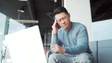 Genç, yorgun, çok çalışan bir ofis çalışanı. Kafasına şiddetli bir baş ağrısı masajı yaparak iş yerinde oturuyor. Sağlıksız portre. Yorgun Asyalı erkek. İş yerindeki sabit bilgisayar stresi yüzünden acı çekiyor.