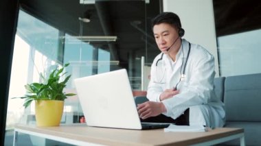 Asyalı erkek doktor hastaya internetten bilgisayar ve kulaklık kullanarak görüntülü görüşme öneriyor. Hekimlerle uzaktan görüşme. Muhabbet eden adam teletıp, tele-sağlık. Ameliyat ofisi