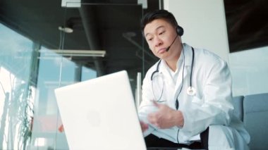 Asyalı erkek doktor hastaya internetten bilgisayar ve kulaklık kullanarak görüntülü görüşme öneriyor. Hekimlerle uzaktan görüşme. Muhabbet eden adam teletıp, tele-sağlık. Ameliyat ofisi