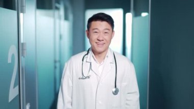 Gülümseyen mutlu Asyalı doktor doktor beyaz bornozuyla modern hastane kliniğinde kameraya bakarak duruyor. Portre erkek doktor terapisti.
