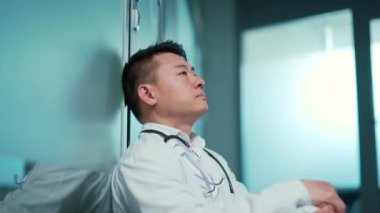 Stresli, yorgun Asyalı doktor koridorda, bir hastanenin içinde oturuyor. Klinikte düşünceli, düşünceli bir doktor. Salgın sırasında steteskopla önlük giyen erkek hasta bakıcısı stresi.