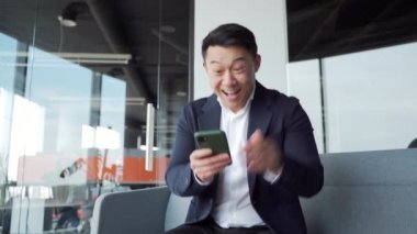 Mutlu Asyalı iş adamı banka kartıyla ödeme yapan online müşteri modern bir ofis satın alma servisinde oturuyor, rezervasyon yapıyor. Memnun olmuş neşeli erkek çalışan cep telefonu akıllı telefonu kullanıyor.