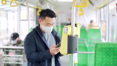 Asyalı adam cep telefonu uygulaması ile toplu taşıma ücretini ödüyor. Yüz maskesi takan erkek yolcu, kontaksız akıllı telefon kullanarak otobüs bileti almış. troleybüs ya da tramvay kullanım uygulaması içinde sür