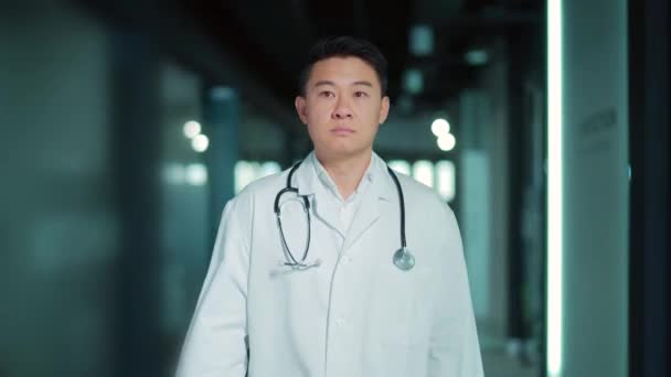 アジア系男性医師が近代病院の廊下を歩いている 屋内ホールに行く深刻な自信のある医療セラピストや外科医の肖像 — ストック動画