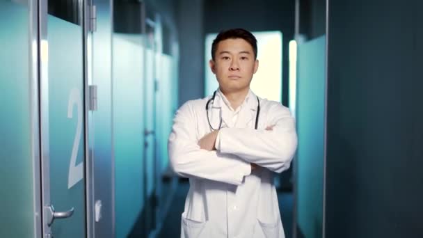肖像画自信的亚洲医生认真地看着摄像机 背景是一个现代化的医院诊所 男医生或男医生双臂交叉地站在室内走廊里 — 图库视频影像