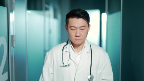 現代の病院の診療所を背景にカメラを真剣に見ている自信に満ちたアジア人医師の肖像画 男性セラピストや医者は彼の腕が交差して室内の廊下に立っている — ストック動画