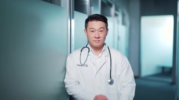 笑顔の幸せな陽気な男性アジアの医師の医療従事者白い衣に手で現代の病院の診療所に立って警察がカメラを見て横切った 頭部撮影ポートレート 男医師セラピスト — ストック動画