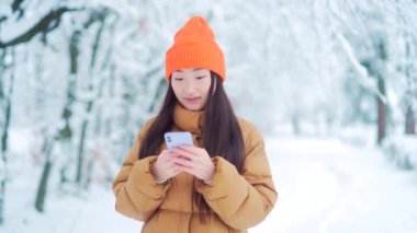 Genç Asyalı bir kadın, parkta bir randevuda, bekliyor, kışın karlı bir günde, online olarak cep telefonunu kullanıyor. Güzellik kızının portresi akıllı telefon kullanarak mesajlaşıyor, kaydırıyor, sohbet ediyor. Açık havada. Soğuk havada