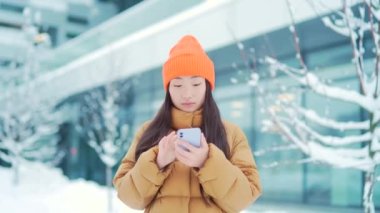 Güzel mutlu Asyalı genç kız öğrenci soğuk havada Kar Şehri caddesinde duruyor. Cep telefonu kullanıyor. Gülümse. Elinde akıllı telefon olan genç bir kadın mesajlaşarak sohbet ediyor. Dışarıda dışarıda.